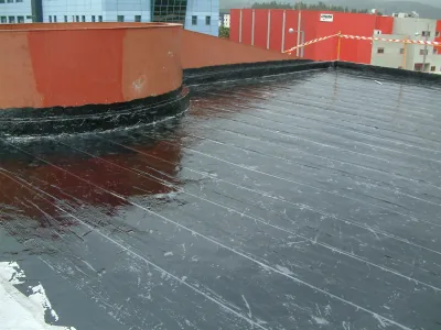 Cubierta con membrana negra brillante que sube por la base de un muro curvo rojizo, con naves al fondo