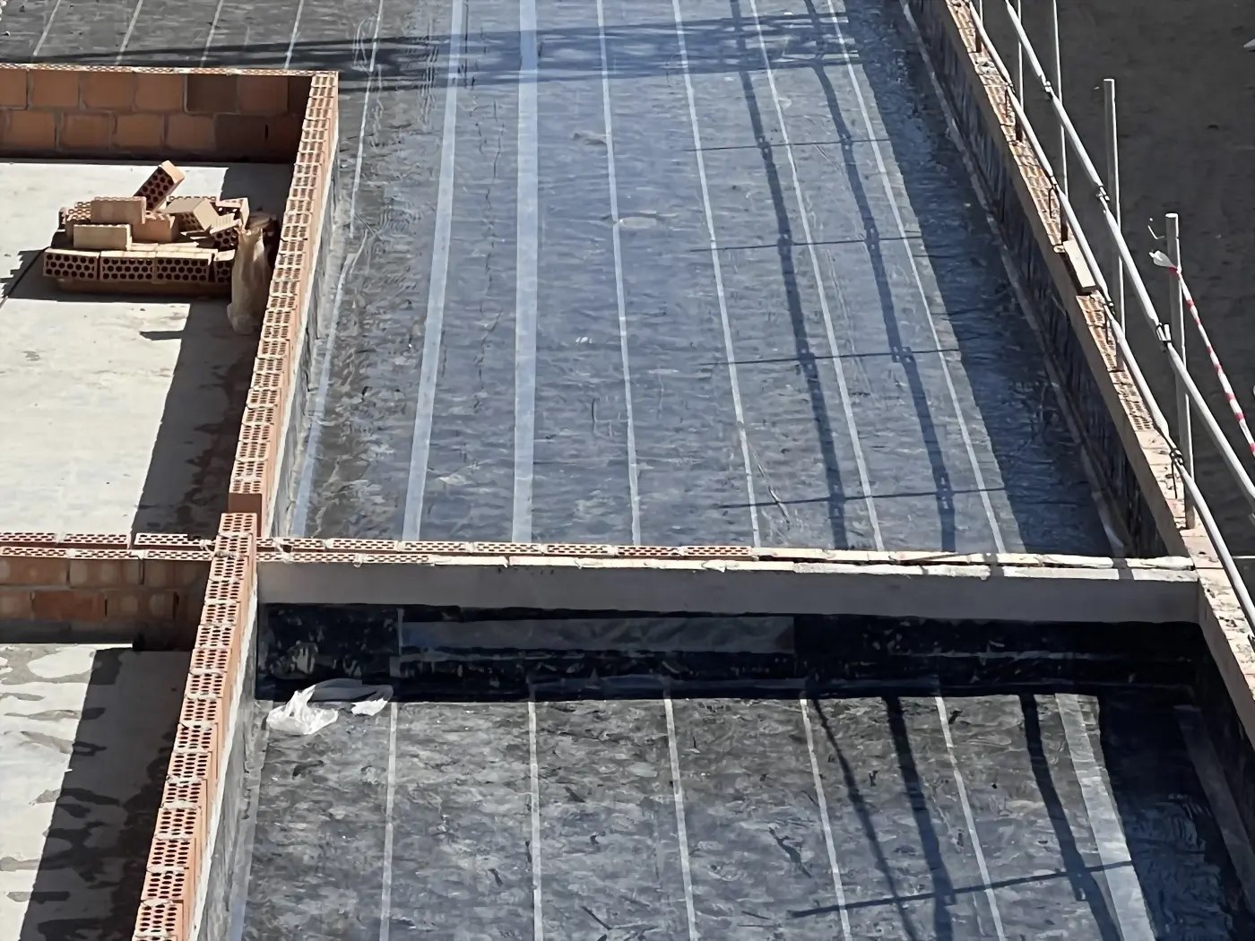Membrana impermeabilizante negra con juntas claras en una terraza en obra, entre murretes de ladrillo y junto a una barandilla