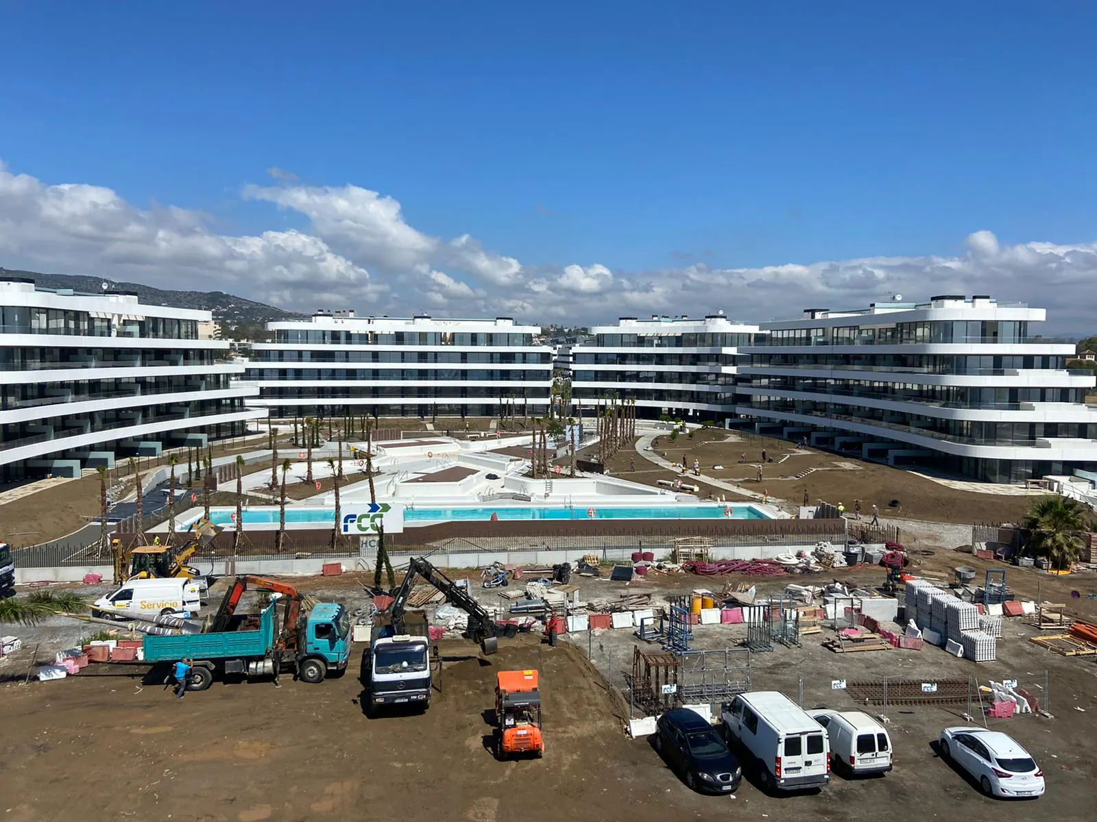 Urbanización de edificios blancos con piscina en fase final de obra, con camiones y maquinaria delante