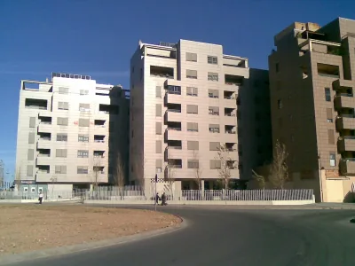 Tres bloques de viviendas de fachada clara junto a una calle curva y un solar, bajo un cielo azul