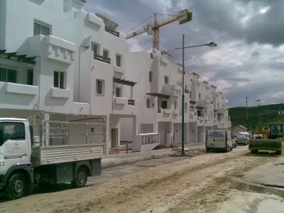Edificios blancos de viviendas en construcción con grúa torre, junto a una calle de tierra con camiones