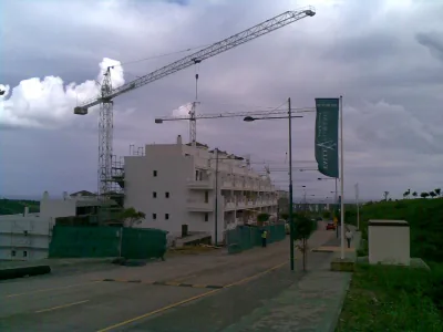 Edificio blanco en construcción con dos grúas torre y una banderola verde junto a una calle, con cielo nublado