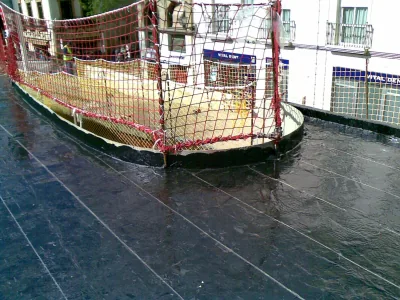 Superficie con membrana negra alrededor de un hueco curvo protegido con red de seguridad roja, junto a locales