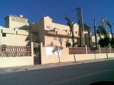Complejo de apartamentos de color crema con muro de celosía rojiza, palmeras y banderas junto a la carretera
