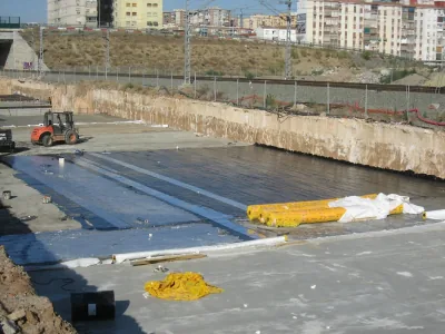 Losa en una trinchera cubierta con membrana negra y franjas claras, con rollos amarillos y un dumper