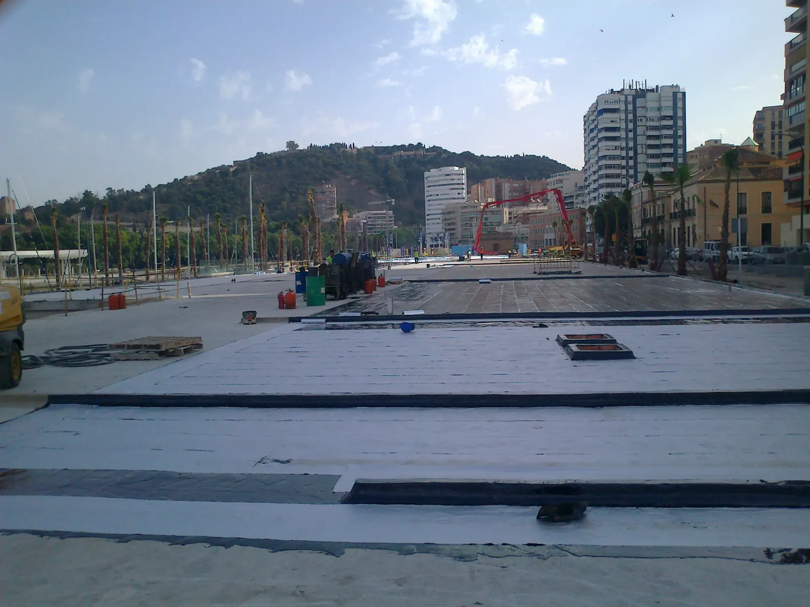 Explanada en obra cubierta con láminas claras y juntas negras, con palmeras, un monte y edificios al fondo