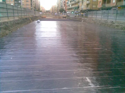 Losa alargada entre edificios cubierta por membrana negra brillante con juntas claras, entre vallas de obra