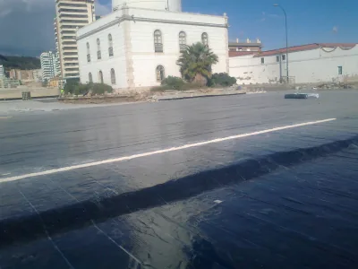 Superficie con membrana negra y una franja clara frente a un edificio blanco con ventanas de arco y una palmera