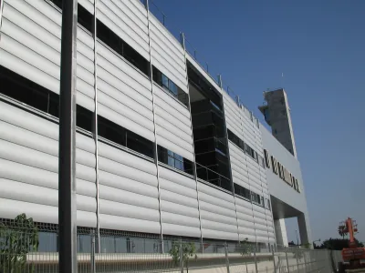 Fachada de un edificio moderno con lamas horizontales blancas y franjas de ventanas oscuras, junto a una torre