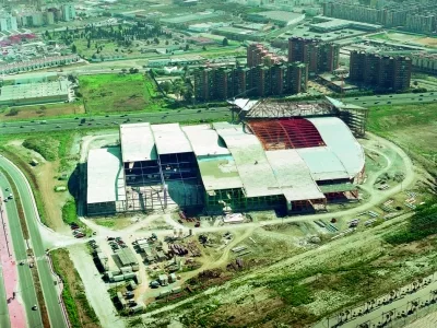 Vista aérea de un gran edificio en construcción con cubiertas blancas y una roja, rodeado de carreteras