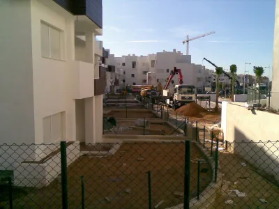 Urbanización de viviendas blancas en obra vista tras una valla metálica, con camiones grúa y tierra